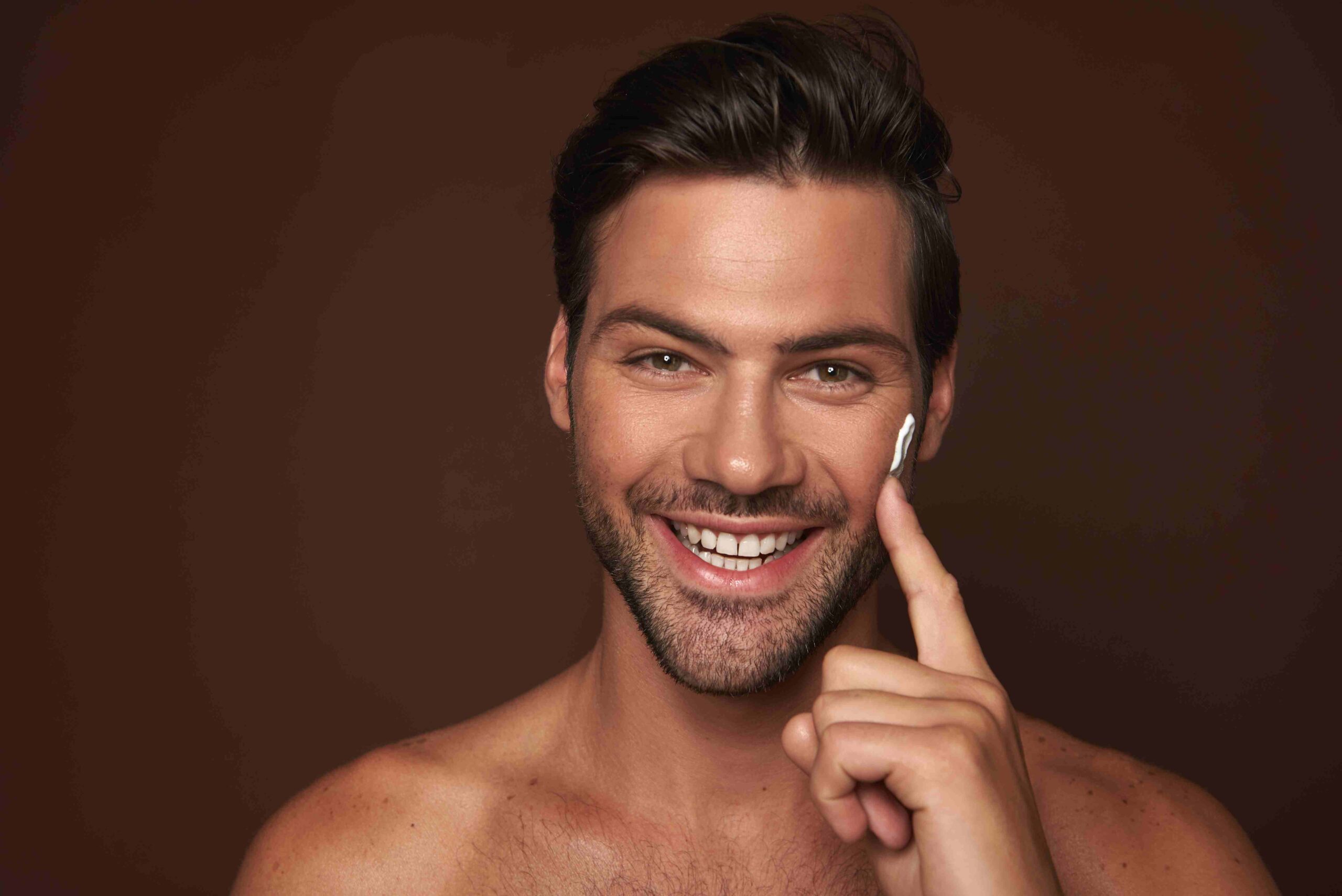 Handsome man with 5 o'clock shadow applying cream to one cheek with index finger