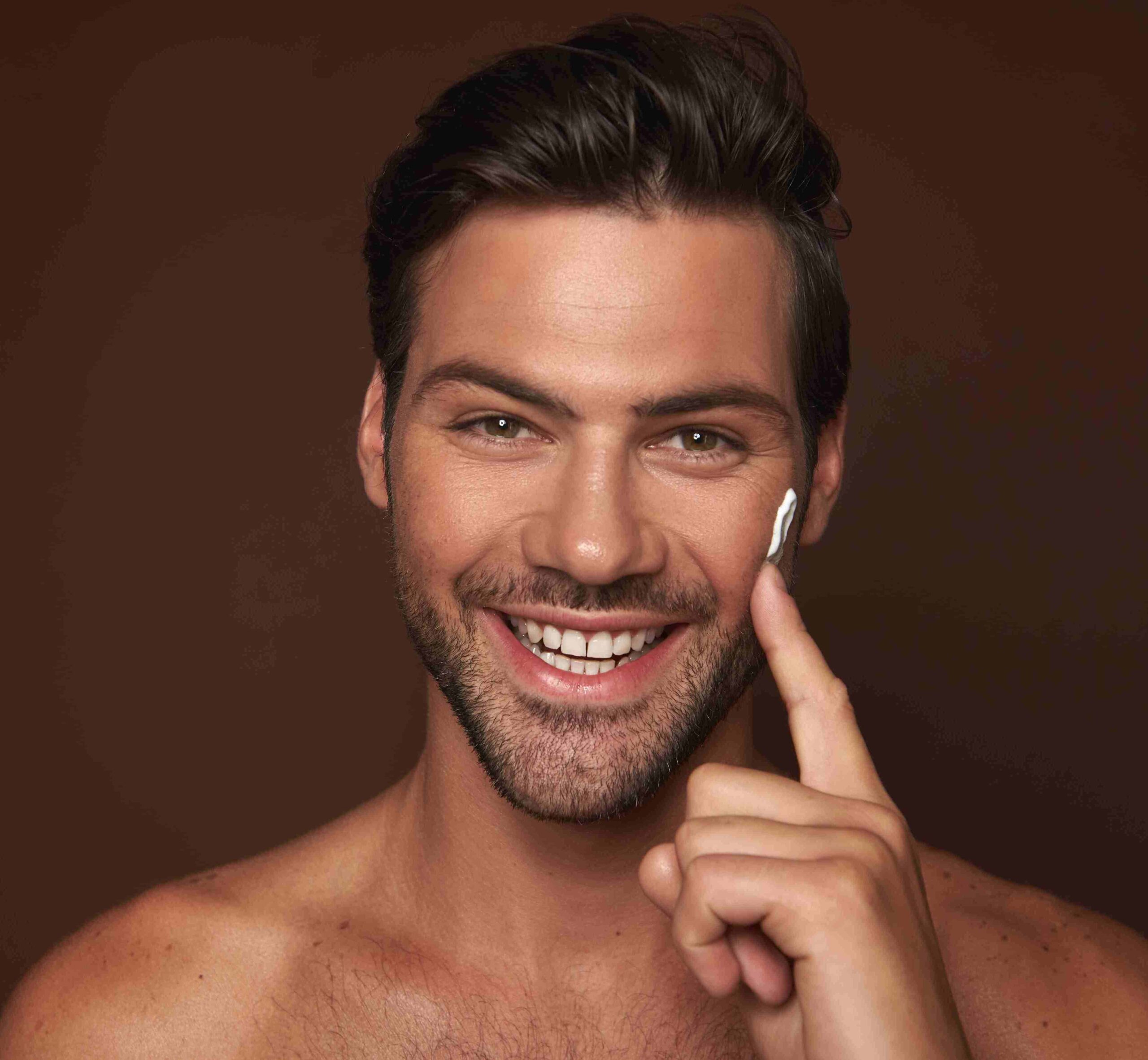 Handsome man with 5 o'clock shadow applying cream to one cheek with index finger